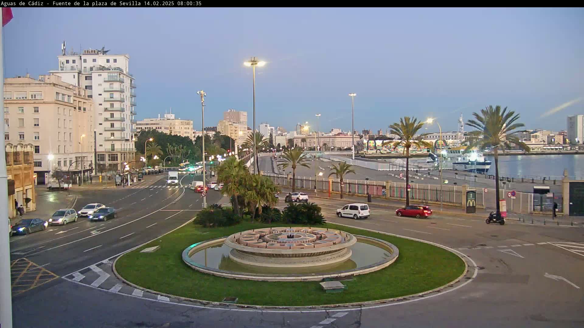 A city street scene at dawn shows a circular fountain in the foreground, with cars driving along a roadway that leads to a harbor, where a ship is docked.