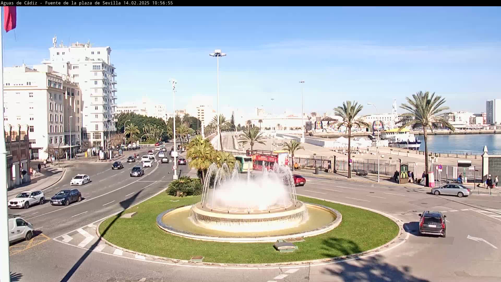 A large fountain is at the center of a traffic circle, with cars driving on the surrounding roads, under a sunny sky, near a waterfront with palm trees and buildings.