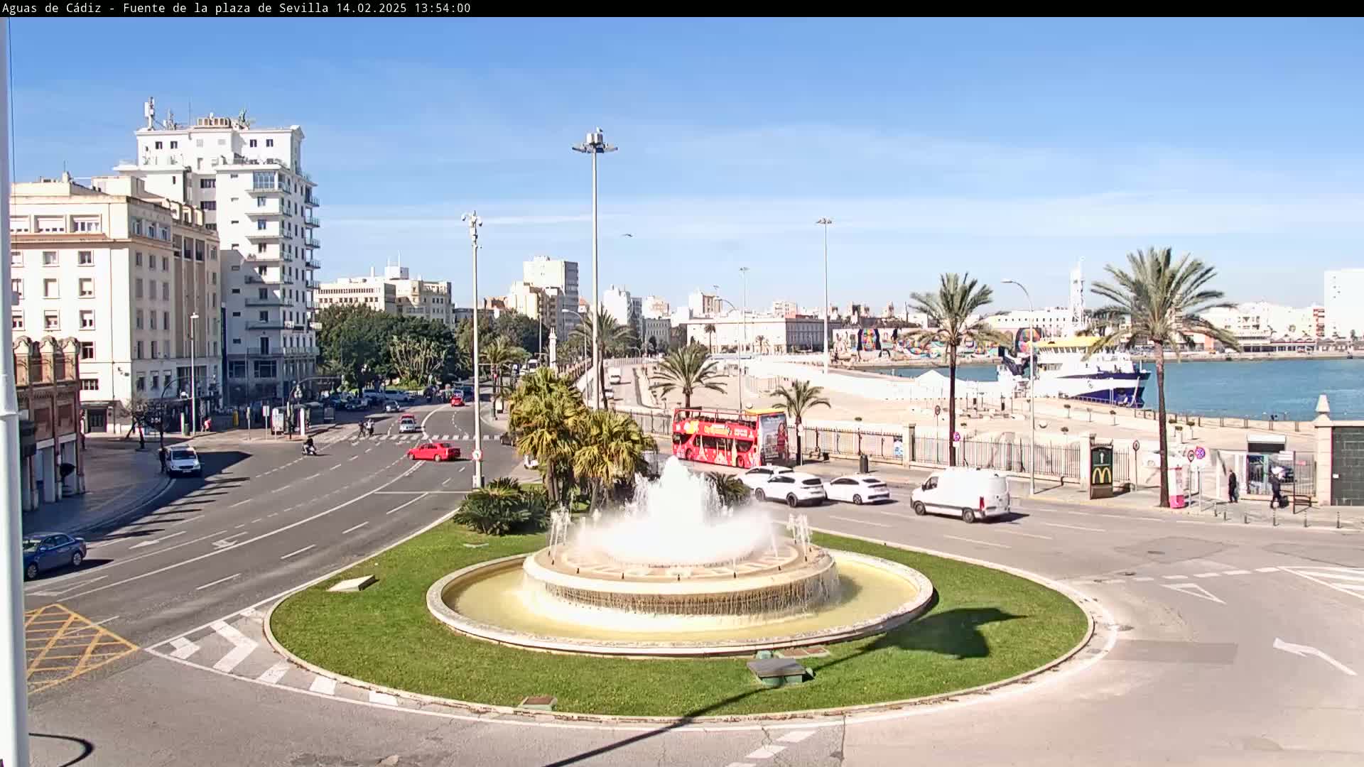 A traffic circle with a fountain in the center features several cars and a double-decker bus, overlooking a harbor with a sunny, clear sky.