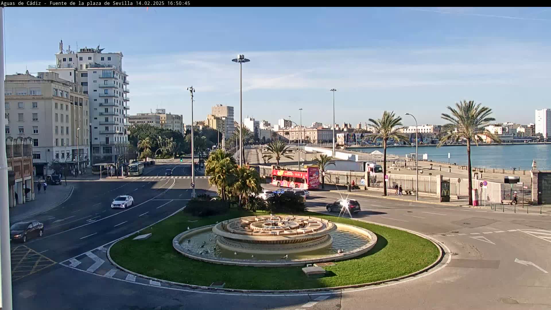A city roundabout with a circular fountain in the center, surrounded by roads, buildings, palm trees, and a body of water in the background under a clear, sunny sky.