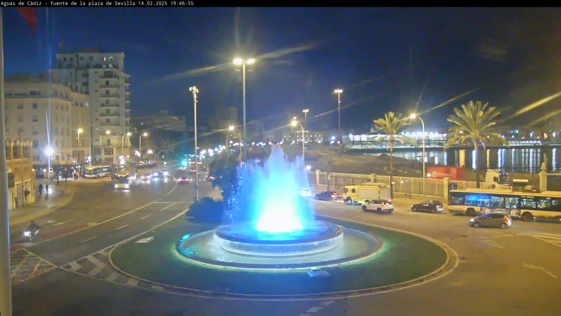 A nighttime view of a city roundabout featuring a brightly lit fountain, with vehicles traveling on the roads surrounding it and buildings and palm trees in the background.