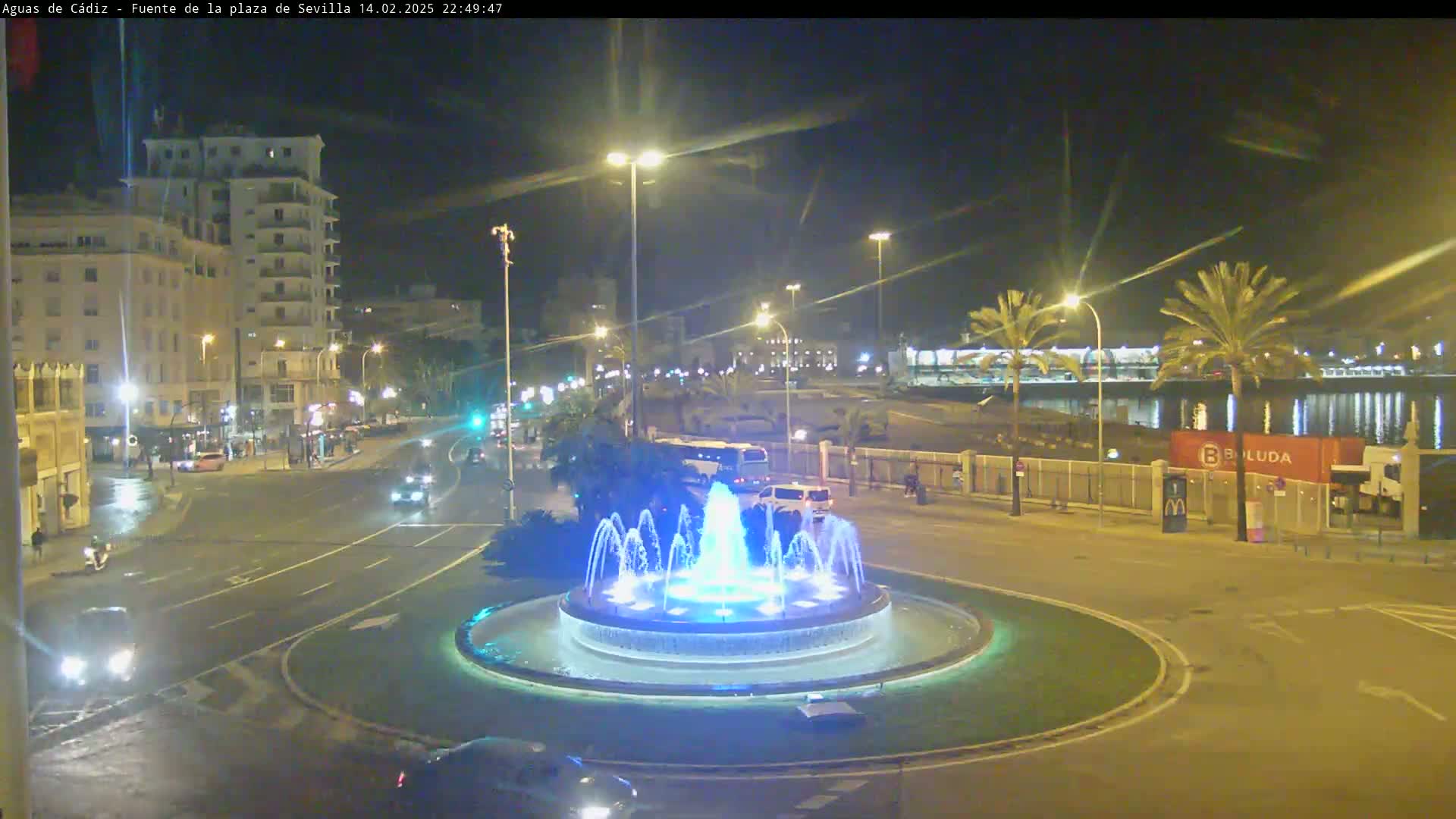 A nighttime view of a city square featuring a circular fountain with blue lights, surrounded by roads with cars, buildings, and palm trees under clear skies.
