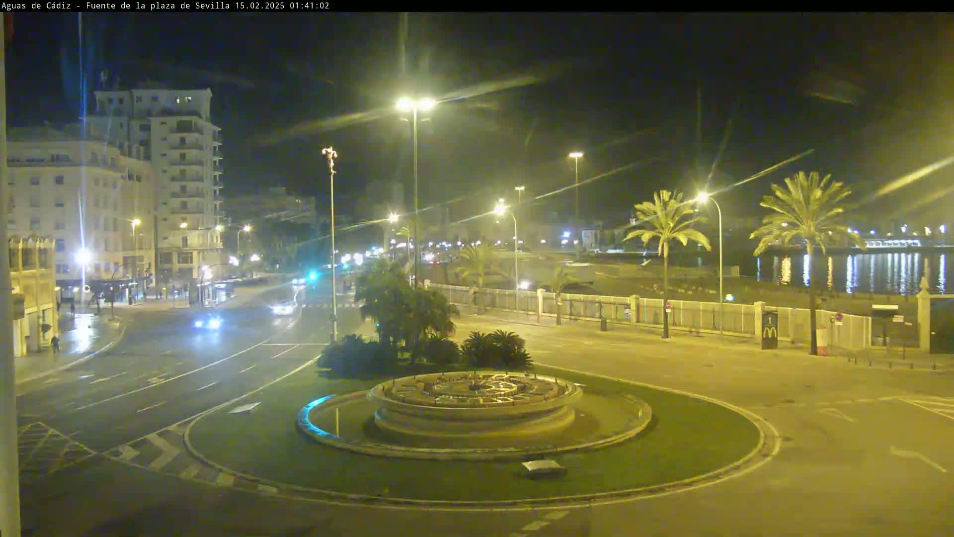 A nighttime view of a city roundabout with a fountain at its center, featuring palm trees, buildings, and some traffic on wet roads.