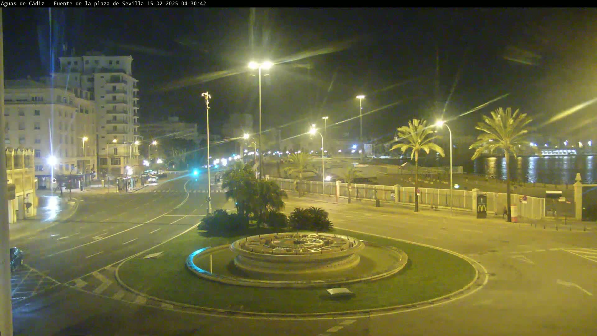A nighttime view of a city roundabout with a fountain in the center, surrounded by palm trees, buildings, and roads, near a waterfront under clear skies.