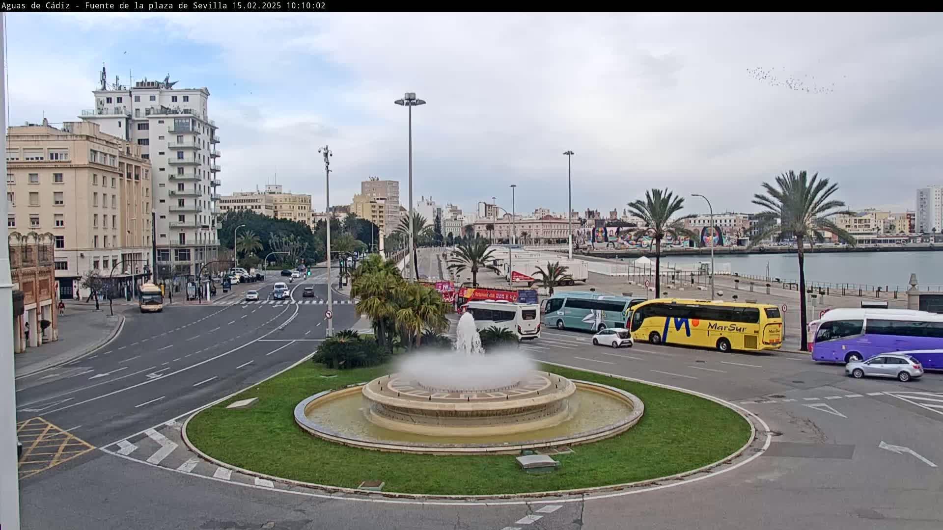 A city square with a fountain in the center, surrounded by roads with cars and buses, under an overcast sky.