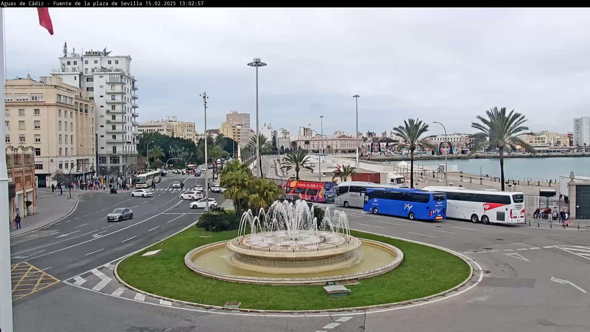 A city square with a fountain at its center, surrounded by buildings, palm trees, and several buses, under an overcast sky.
