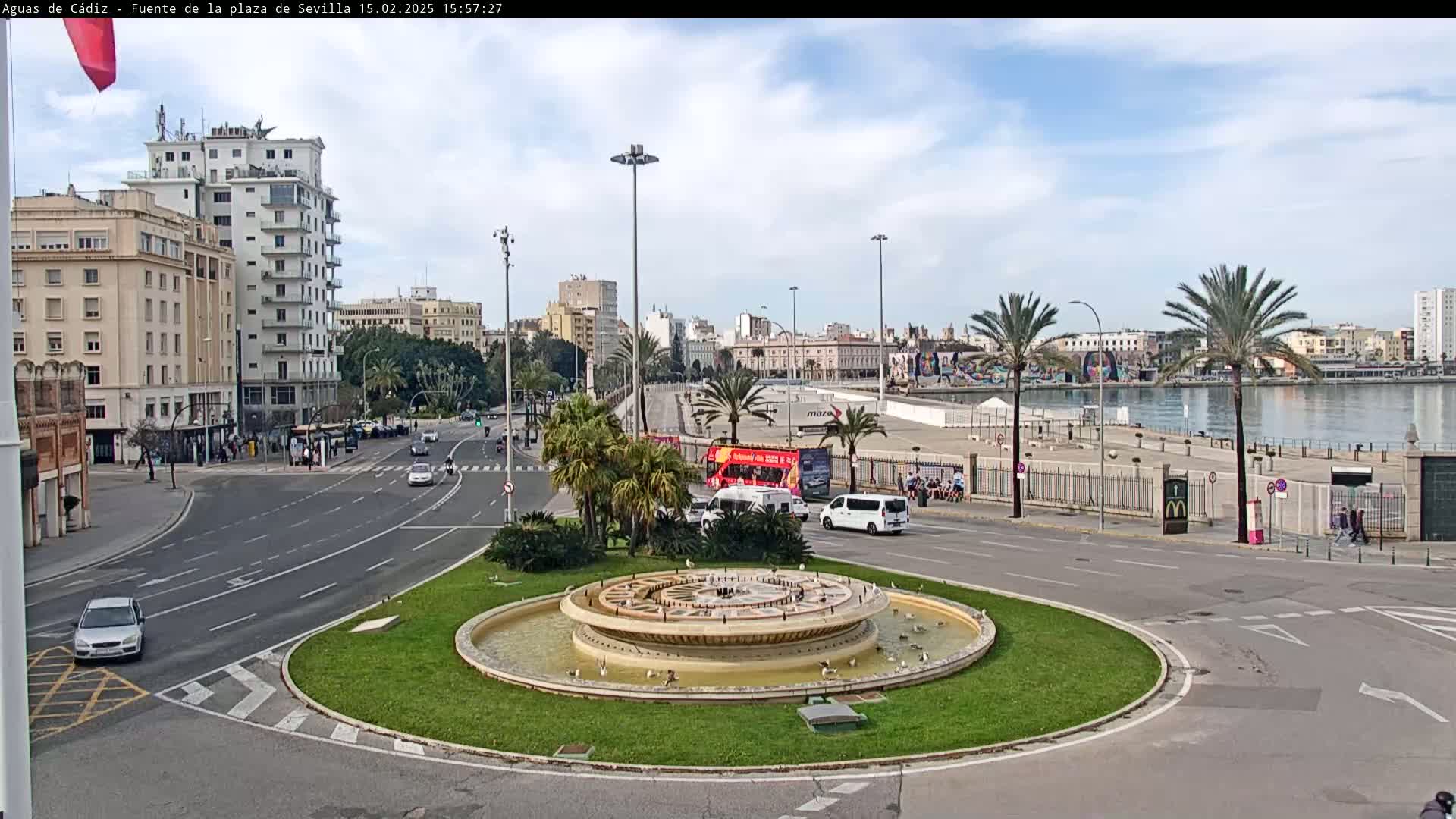 A circular fountain sits in the center of a traffic circle, with city buildings and a waterfront visible in the background under a partly cloudy sky.