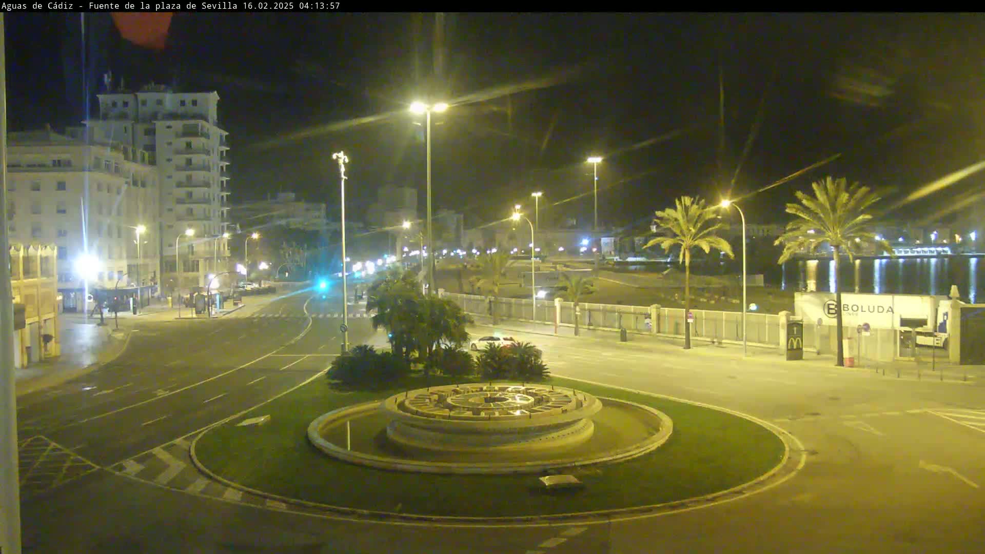 A nighttime view of a circular fountain in a city square, with palm trees and buildings lining a road under bright streetlights.