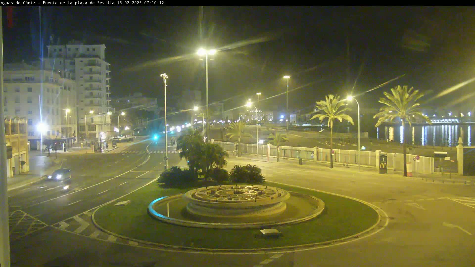 A nighttime view of a circular traffic area with a fountain at its center, surrounded by palm trees, buildings, and a waterfront.