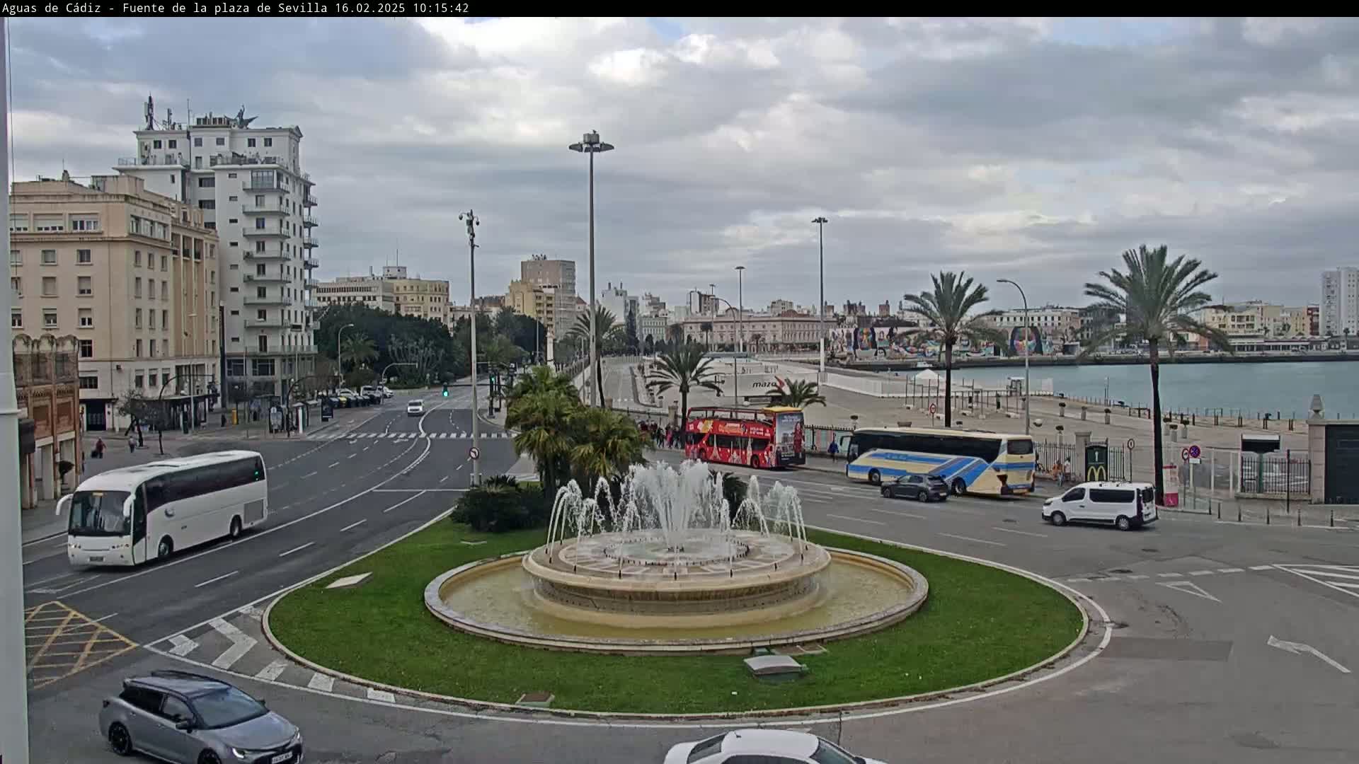 A circular fountain is in the center of a traffic circle with several buses and cars near a waterfront city under an overcast sky.