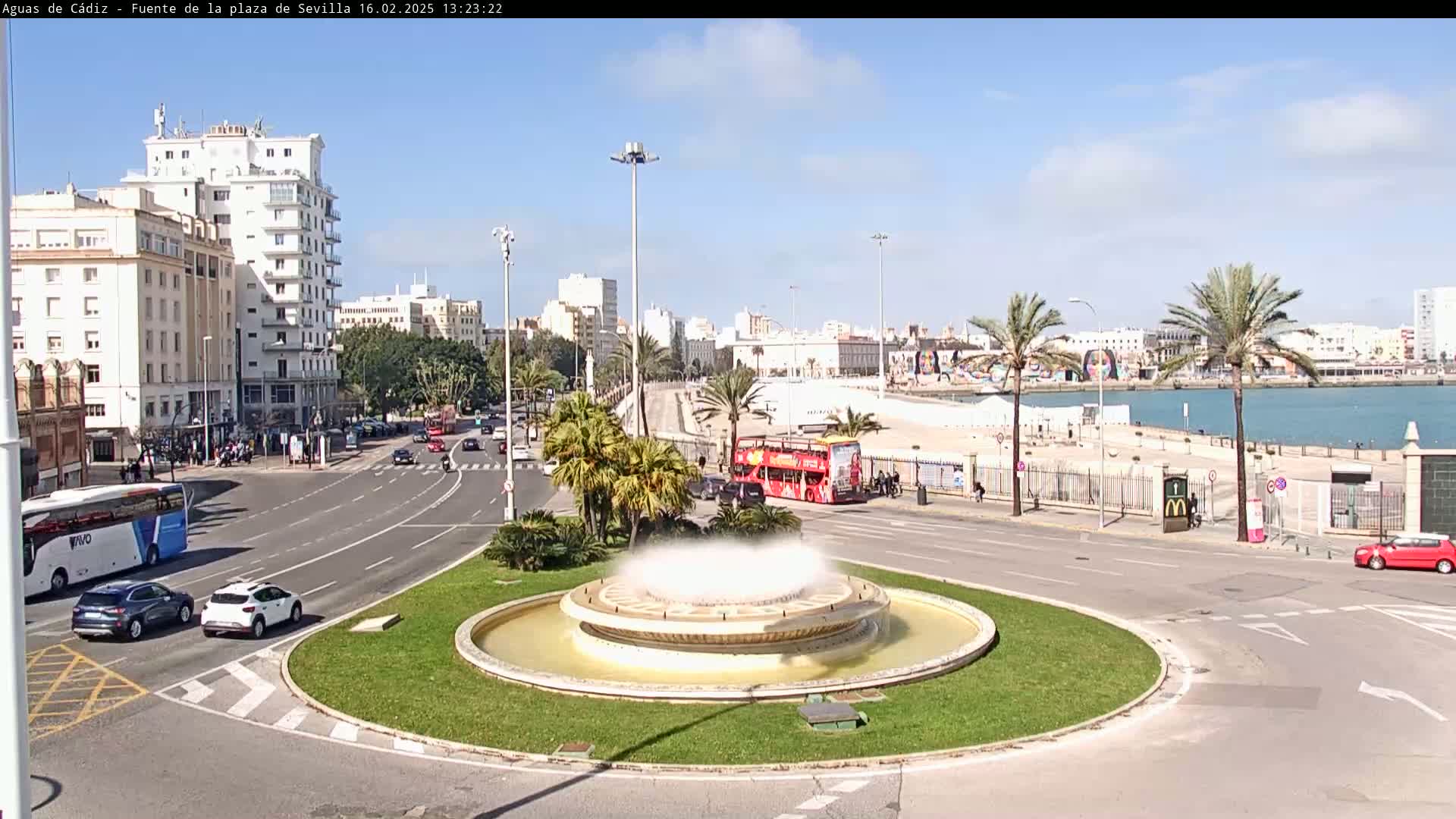 A circular fountain in a city square sprays water on a sunny day, with a waterfront and buildings in the background.