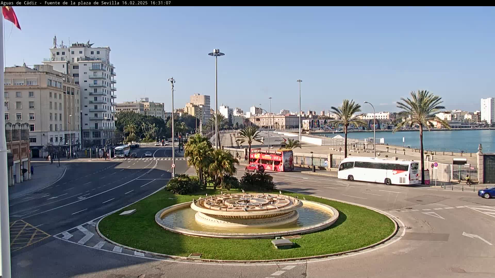 A circular fountain sits in the center of a plaza, with city buildings, palm trees, buses, and a body of water visible in the background under a clear, sunny sky.