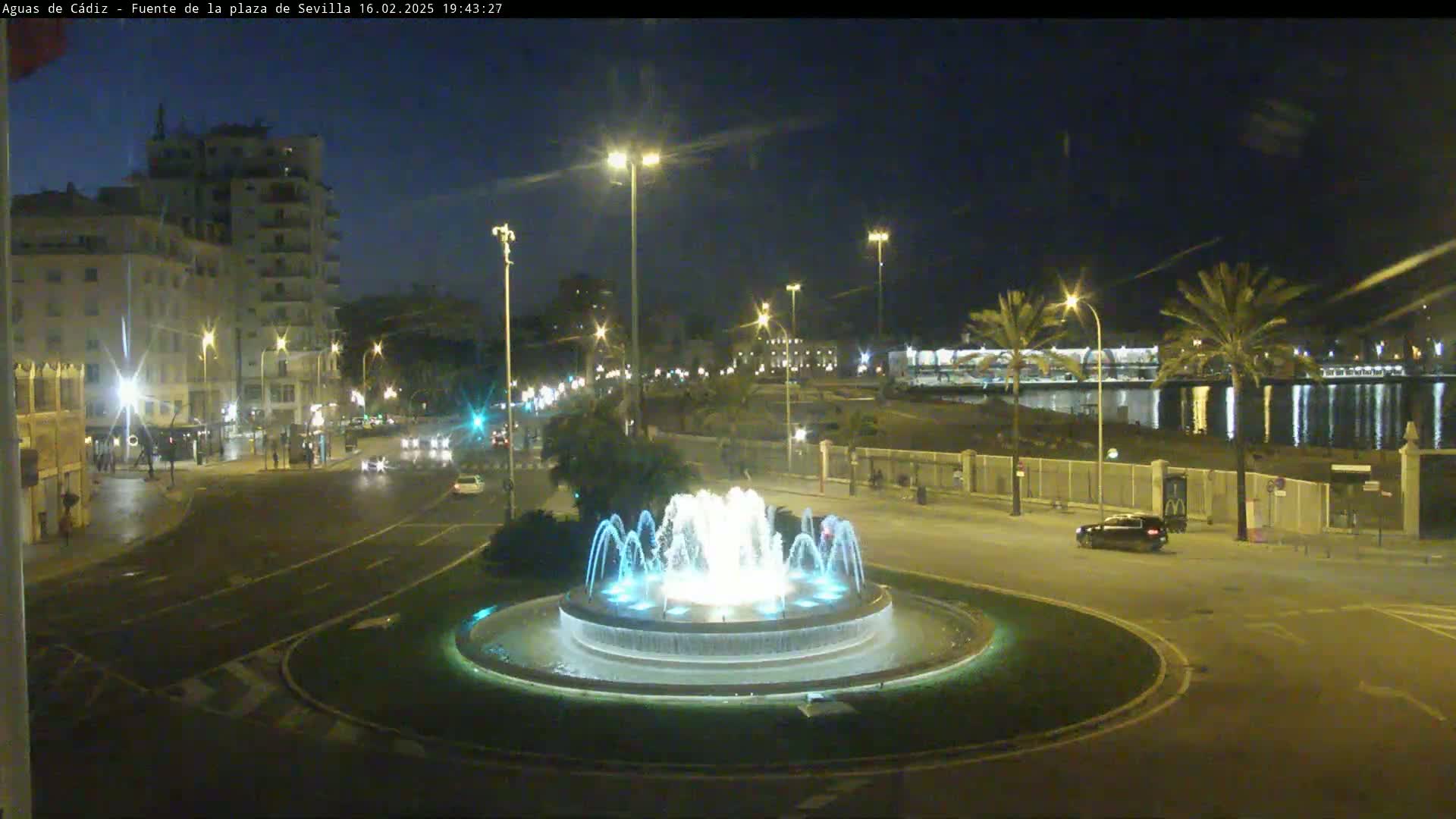 A nighttime view of a city street with a circular fountain at its center, illuminated by streetlights, and bordered by buildings and palm trees under a clear sky.