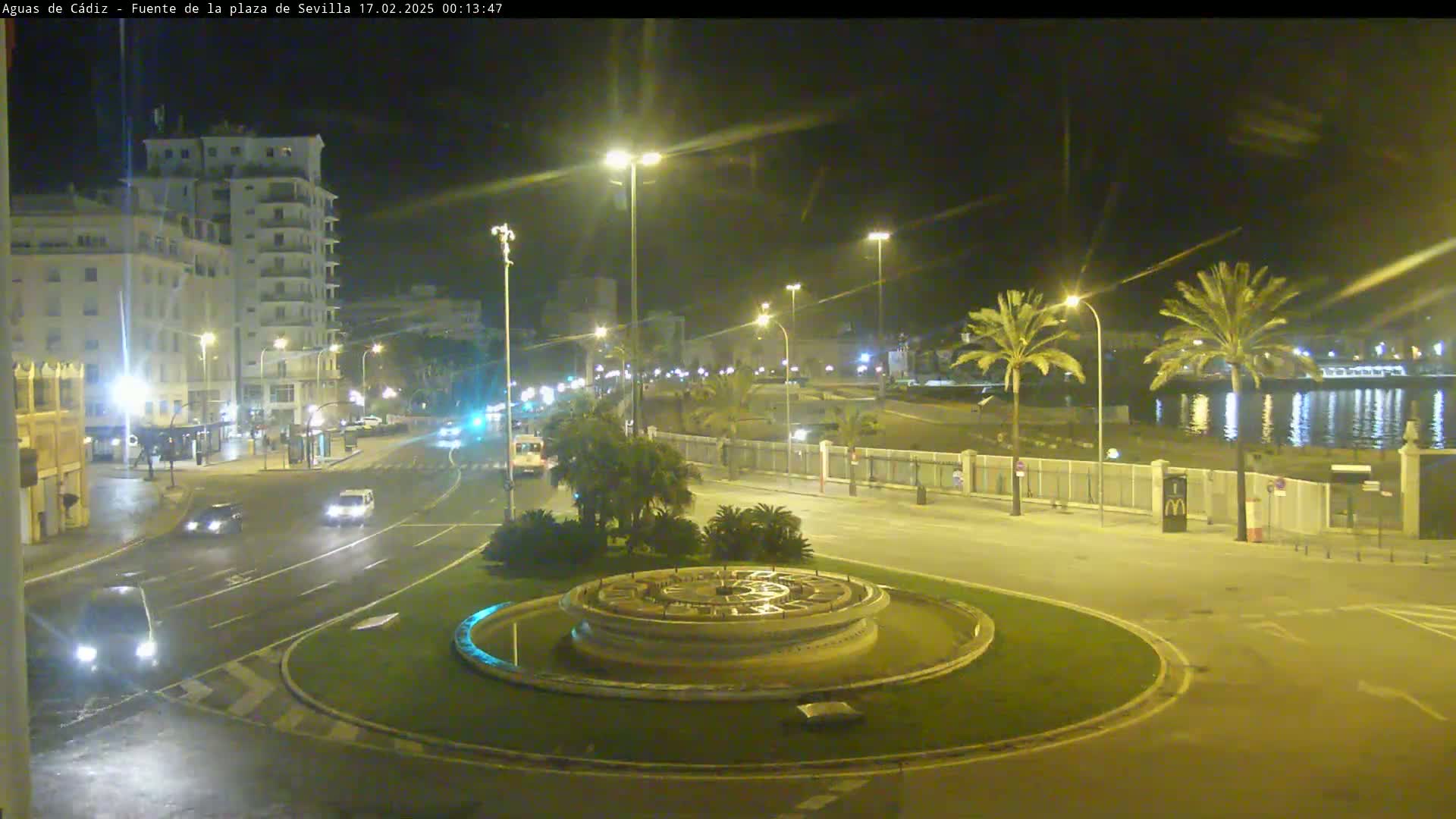 A nighttime view of a city roundabout with a fountain at its center, bordered by palm trees, buildings, and a waterfront, with cars driving on the surrounding roads.