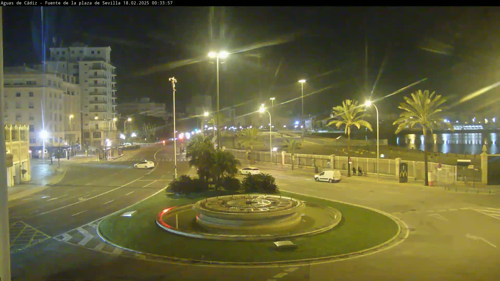 A nighttime view of a city roundabout with a circular fountain at its center, illuminated by streetlights, overlooking a calm body of water and palm trees under a clear sky.
