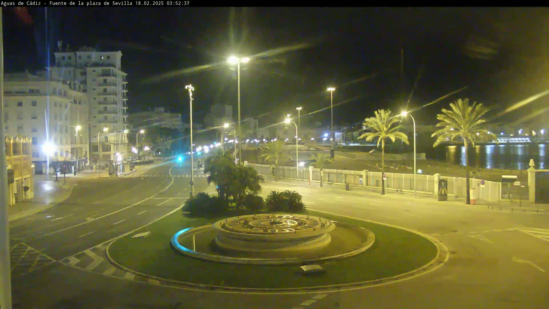 A nighttime view of a city roundabout with a fountain at its center, bordered by palm trees, buildings, and a calm body of water in the background.