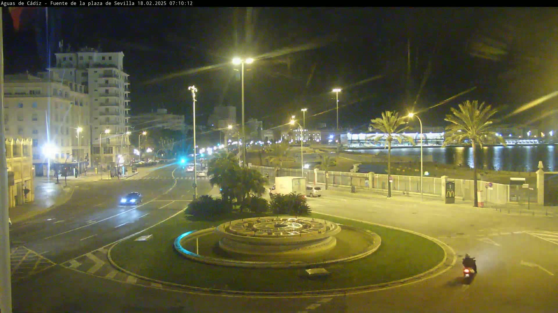 A nighttime view of a city roundabout with a circular fountain at its center, near a body of water and under clear skies.