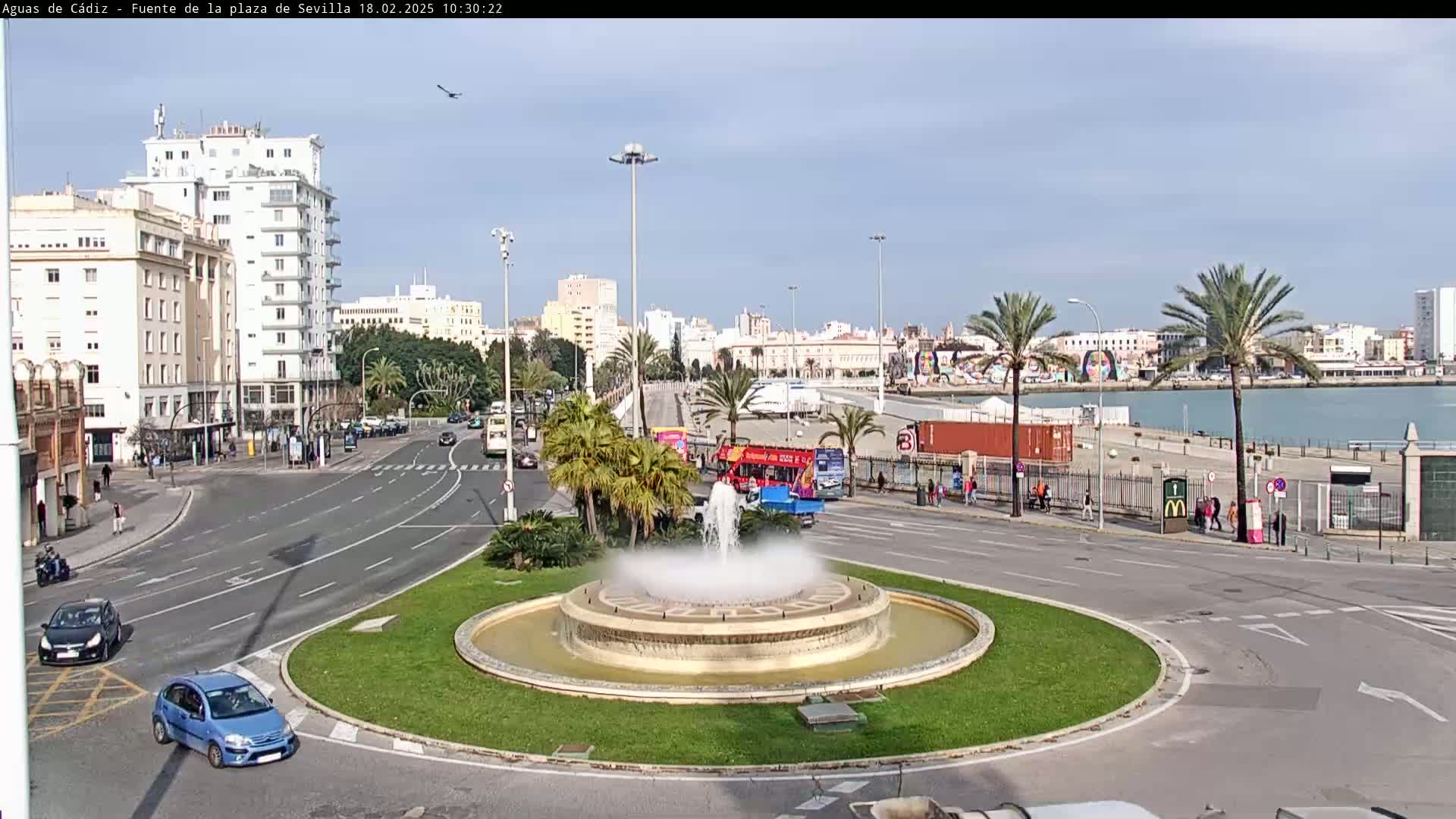 A city fountain is the centerpiece of a roundabout with palm trees, buildings, and a waterfront visible in the background on a sunny day.