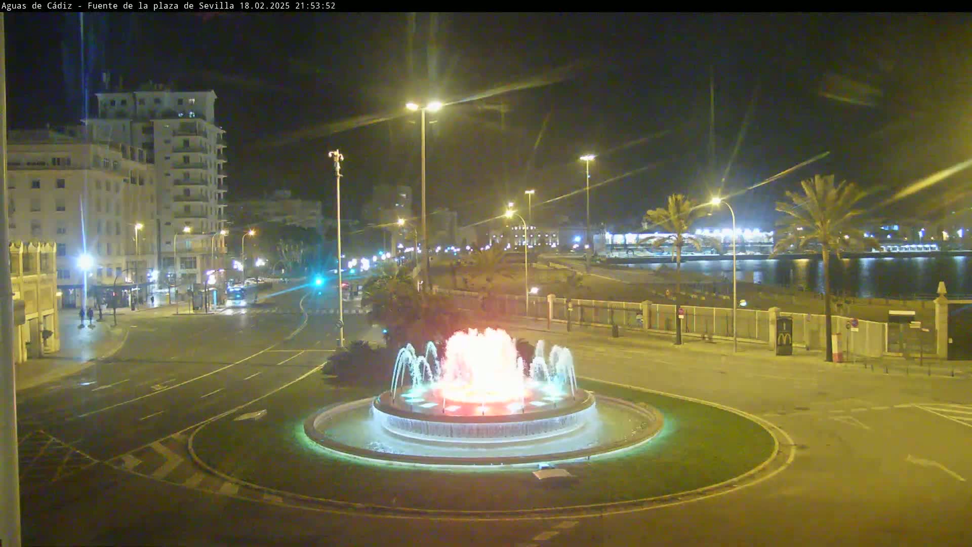 A large illuminated fountain is in a city square at night, with buildings and a waterfront visible in the background.