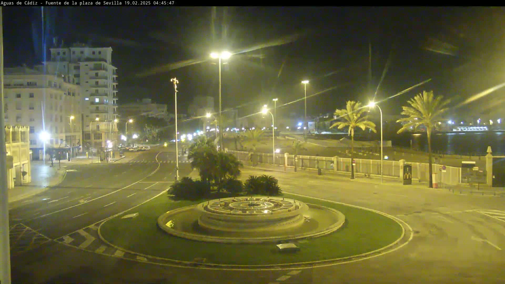A nighttime view of a city roundabout with a circular fountain at its center, illuminated by streetlights, with palm trees and a building visible in the background.