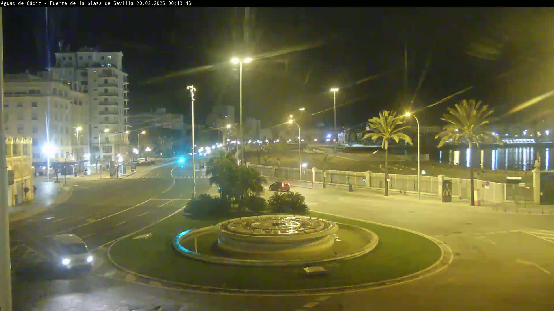 A nighttime view of a city roundabout with a fountain at its center, illuminated by streetlights, showing a few vehicles and palm trees along a waterfront.