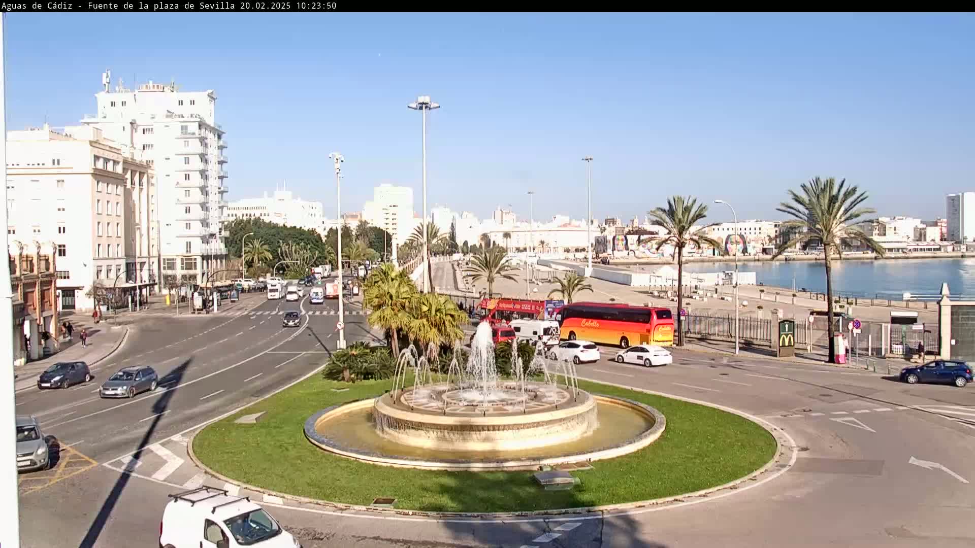 A circular fountain is at the center of a traffic circle, surrounded by palm trees, buildings, and cars, under a clear blue sky.