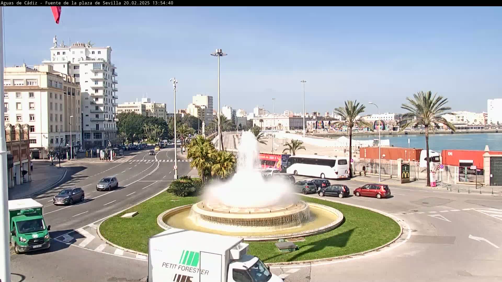 A large fountain sprays water in a city square, with cars driving on surrounding roads and buildings, including a high-rise, visible in the background under a clear sky.