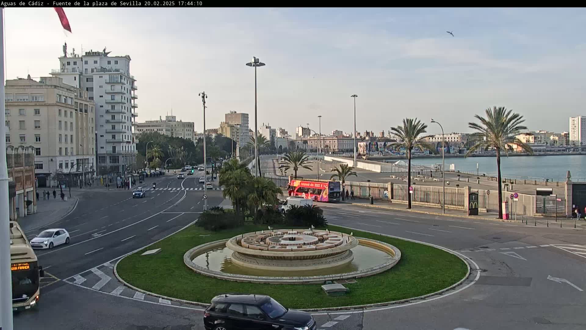 A city roundabout with a central fountain is shown, with a double-decker bus and several cars visible on roads leading to the waterfront under a mostly clear sky.