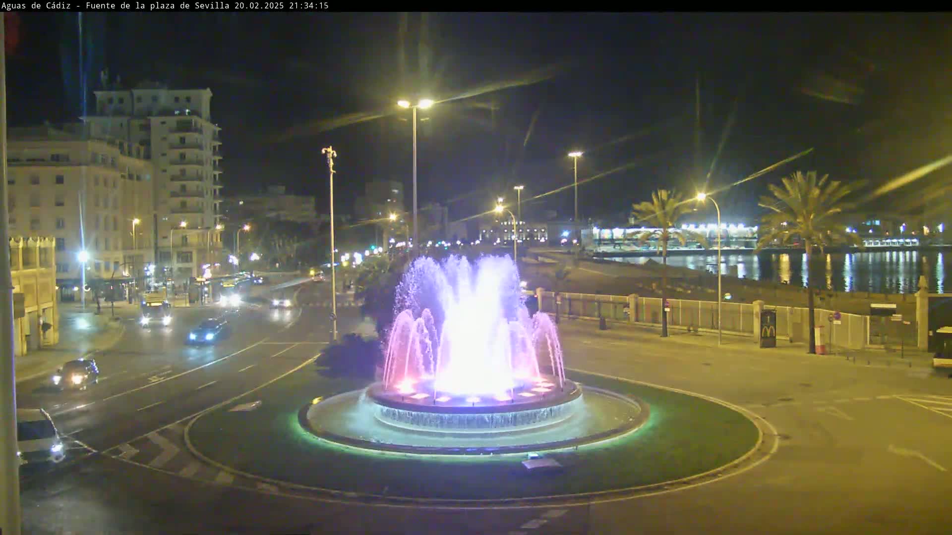 A brightly lit fountain in a roundabout at night, surrounded by cars and buildings, with a calm body of water visible in the background.