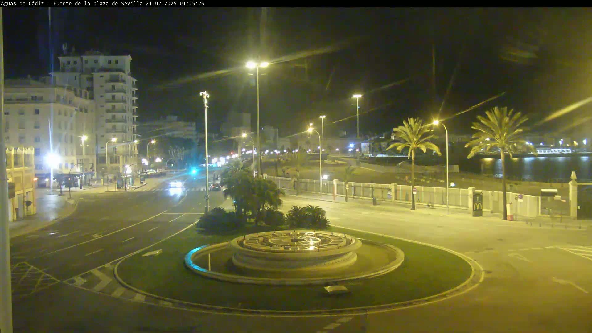 A nighttime view of a city roundabout with a circular fountain at its center, flanked by palm trees, buildings, and roadways, under clear night skies.