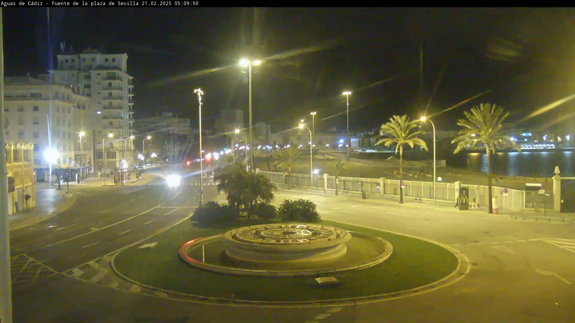 A nighttime view of a city roundabout with a circular fountain in the center,  palm trees lining a waterfront, and buildings illuminated by streetlights.