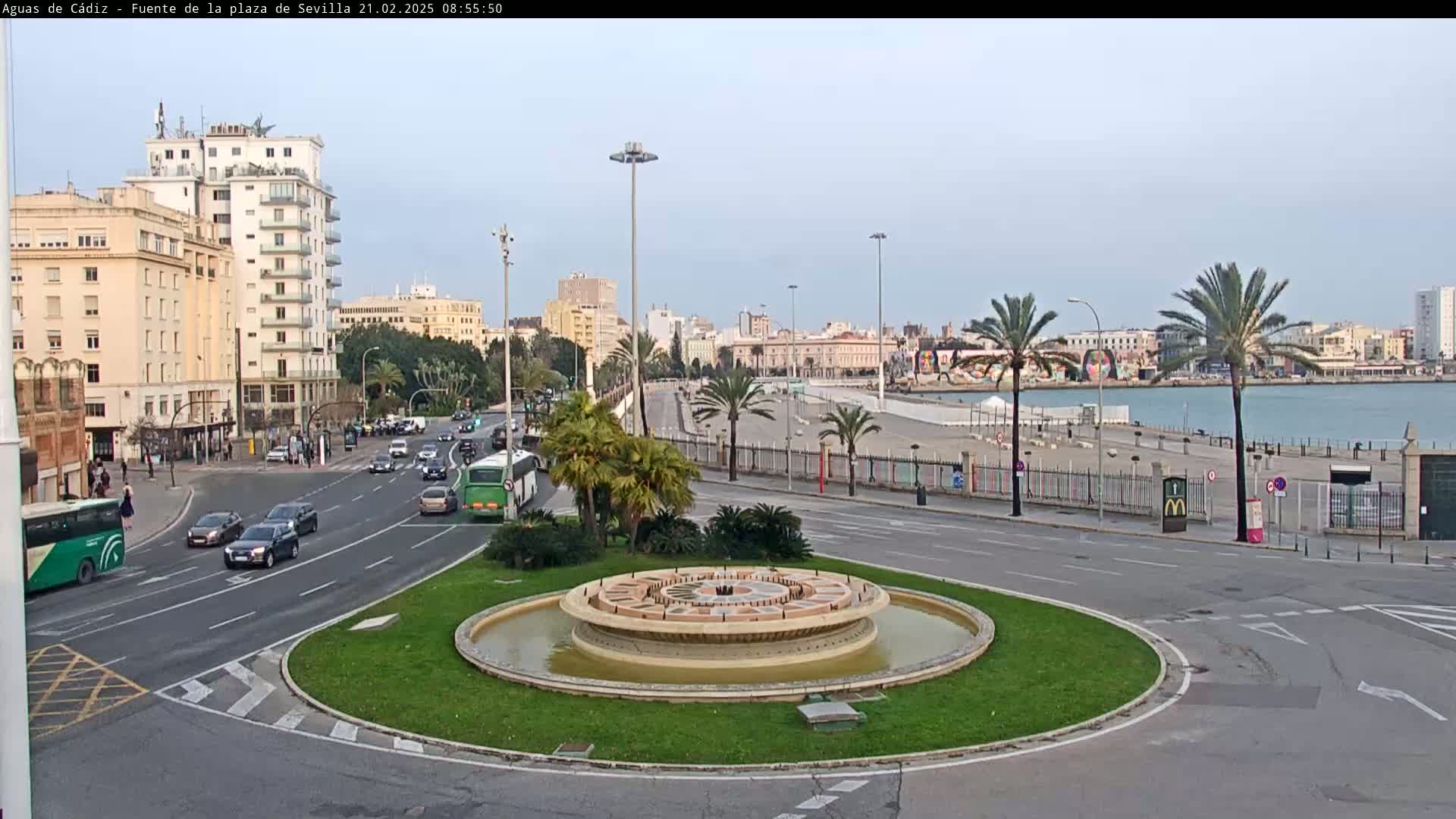 A city roundabout with a circular fountain in the center, overlooking a calm body of water and a waterfront promenade under a mostly clear sky.