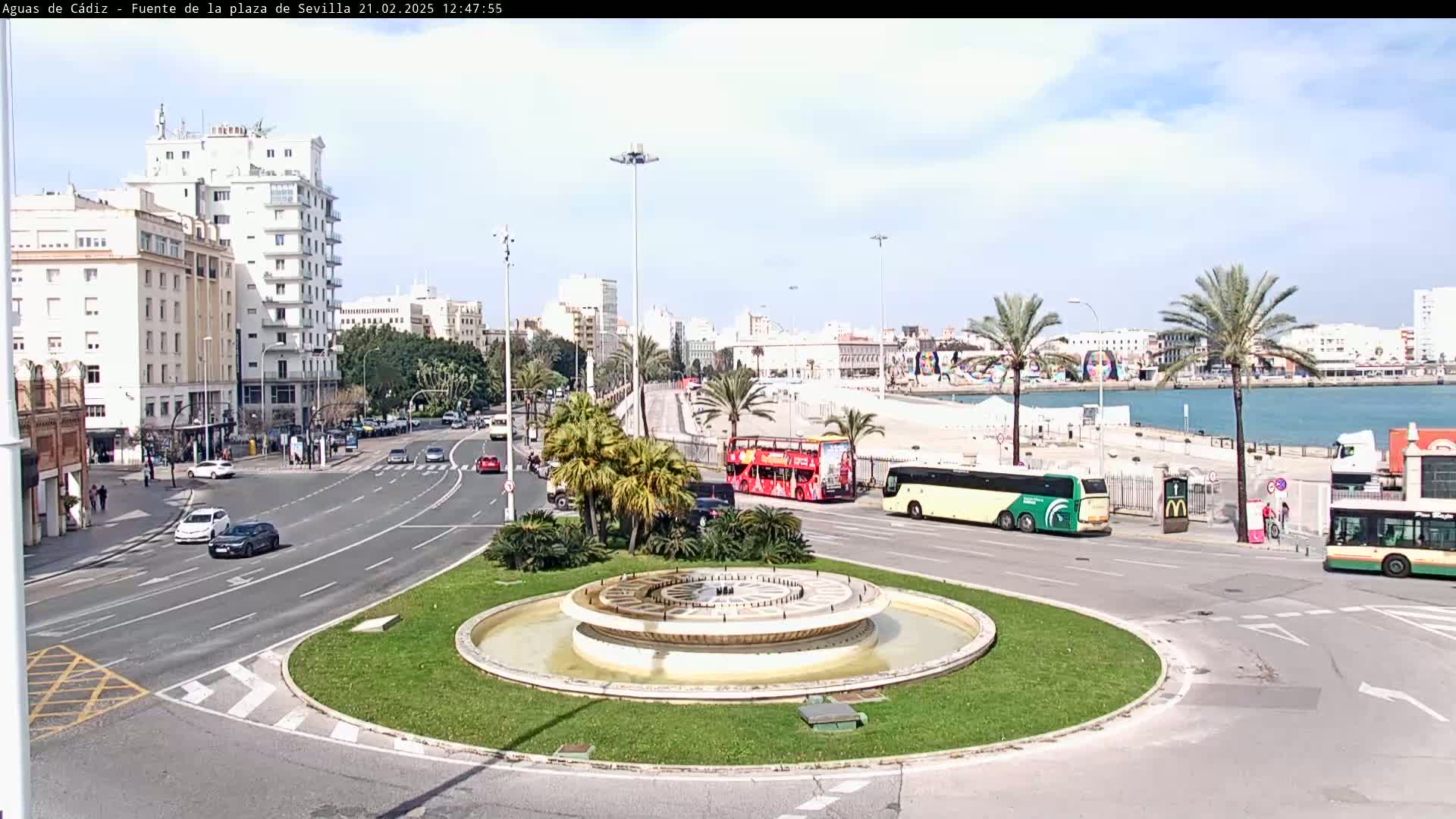A circular fountain is at the center of a traffic circle, with several cars, buses, and palm trees surrounding it, all under a partly cloudy sky.
