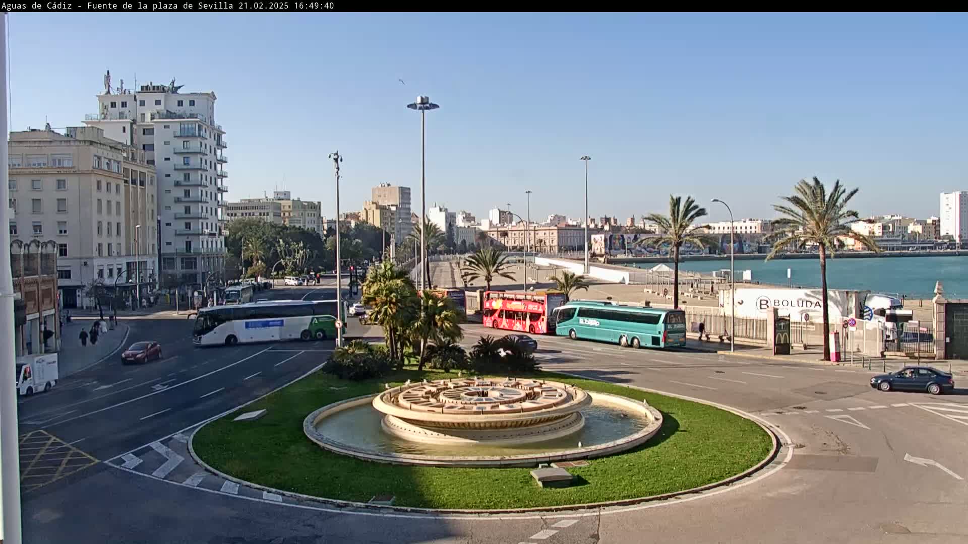A circular fountain sits in the center of a traffic circle, surrounded by buildings, buses, cars, and palm trees, under a clear, sunny sky near a body of water.