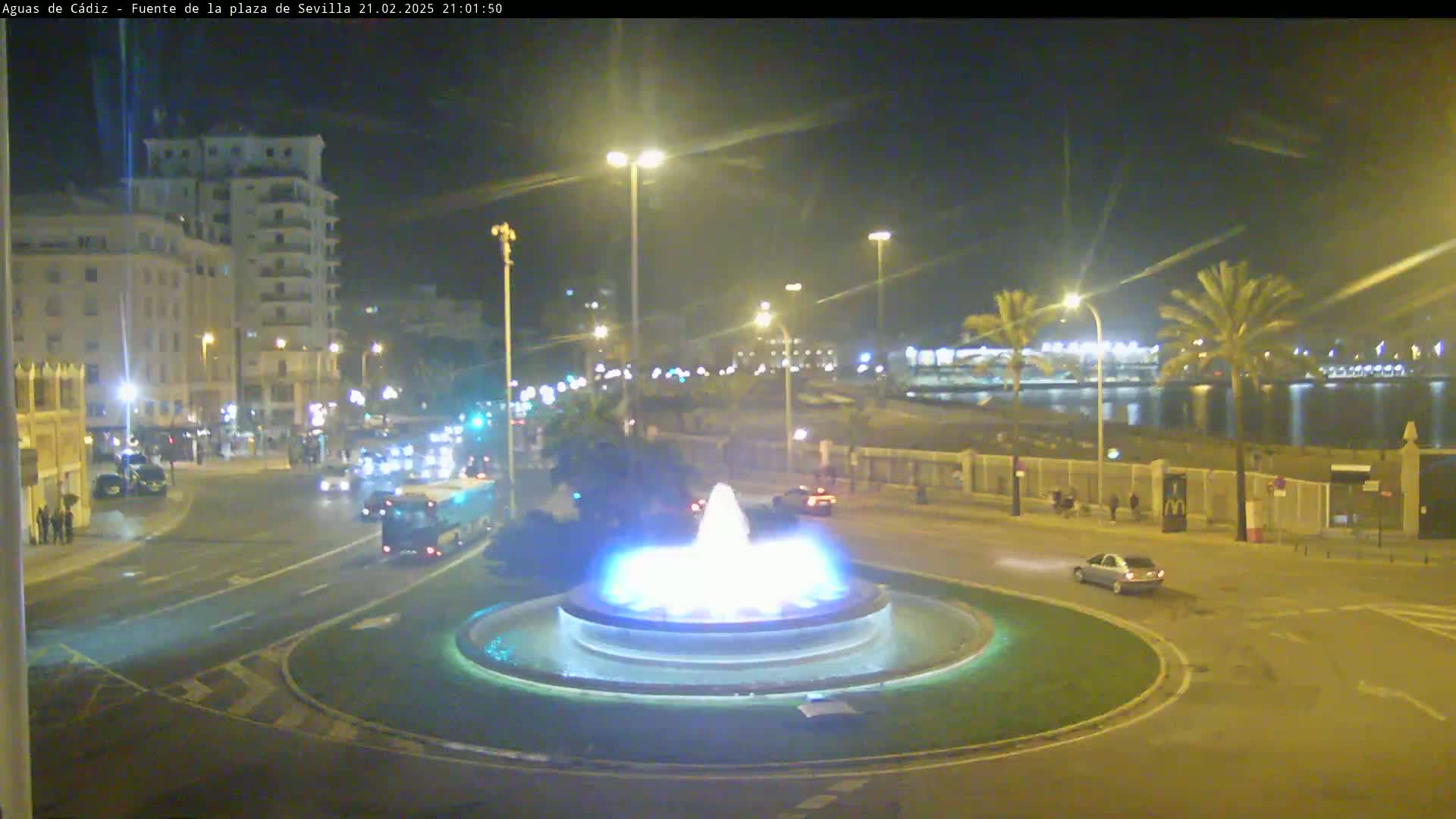 A nighttime view of a city roundabout featuring a lit fountain, with cars driving on the road and buildings and palm trees in the background.