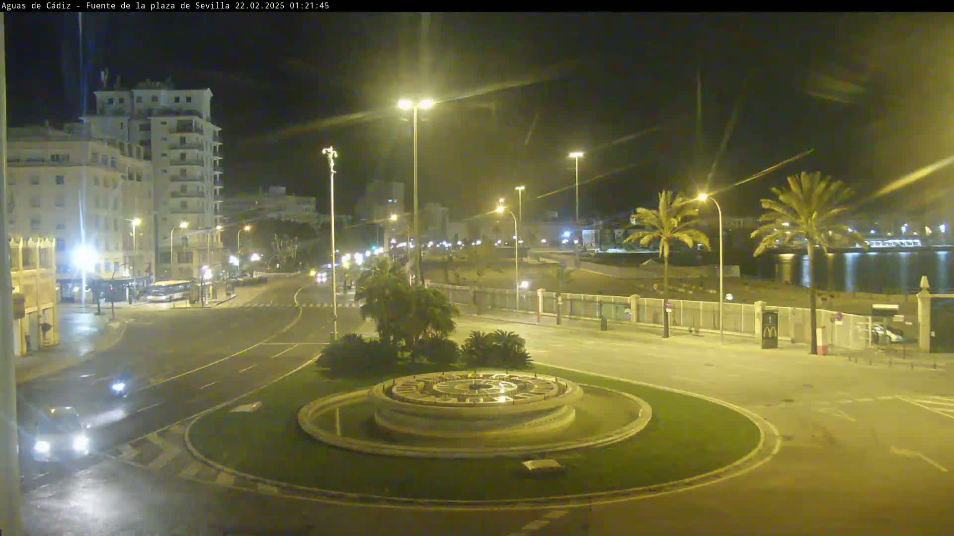 A nighttime view of a circular plaza with a fountain in the center, surrounded by palm trees, buildings, and a roadway with sparse traffic.