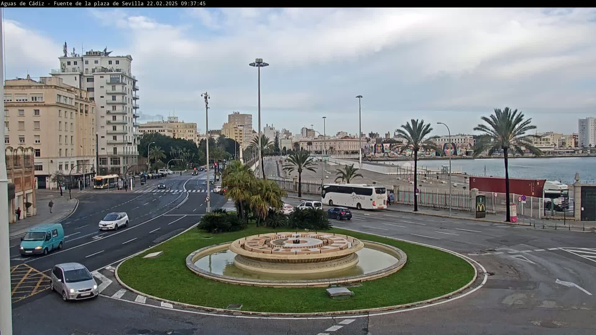 A city roundabout with a circular fountain in the center, surrounded by buildings, palm trees, and cars, on an overcast day.
