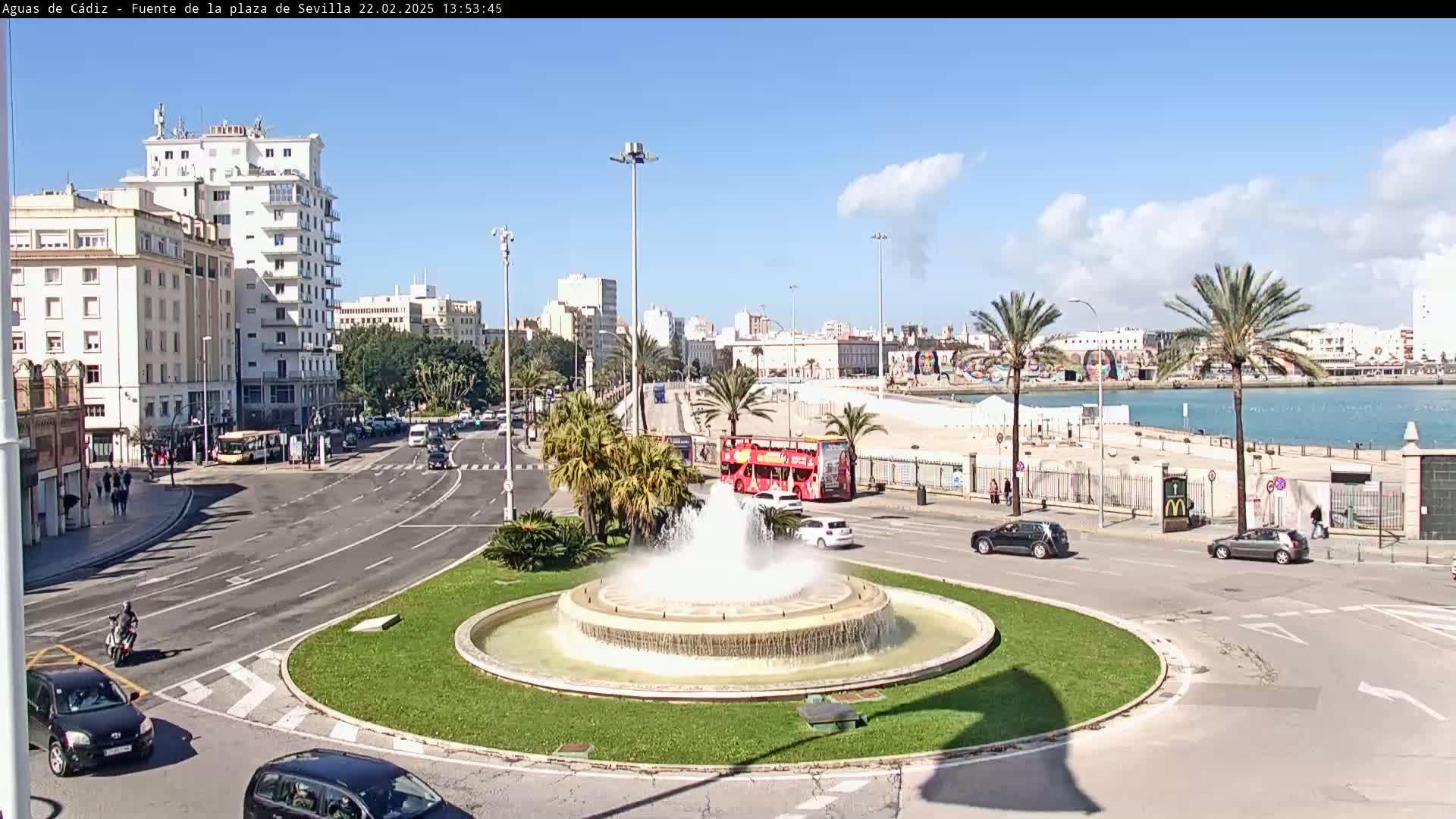 A city street scene on a sunny day shows a circular traffic area with a fountain in the center, cars driving around it, and buildings and palm trees in the background near a body of water.