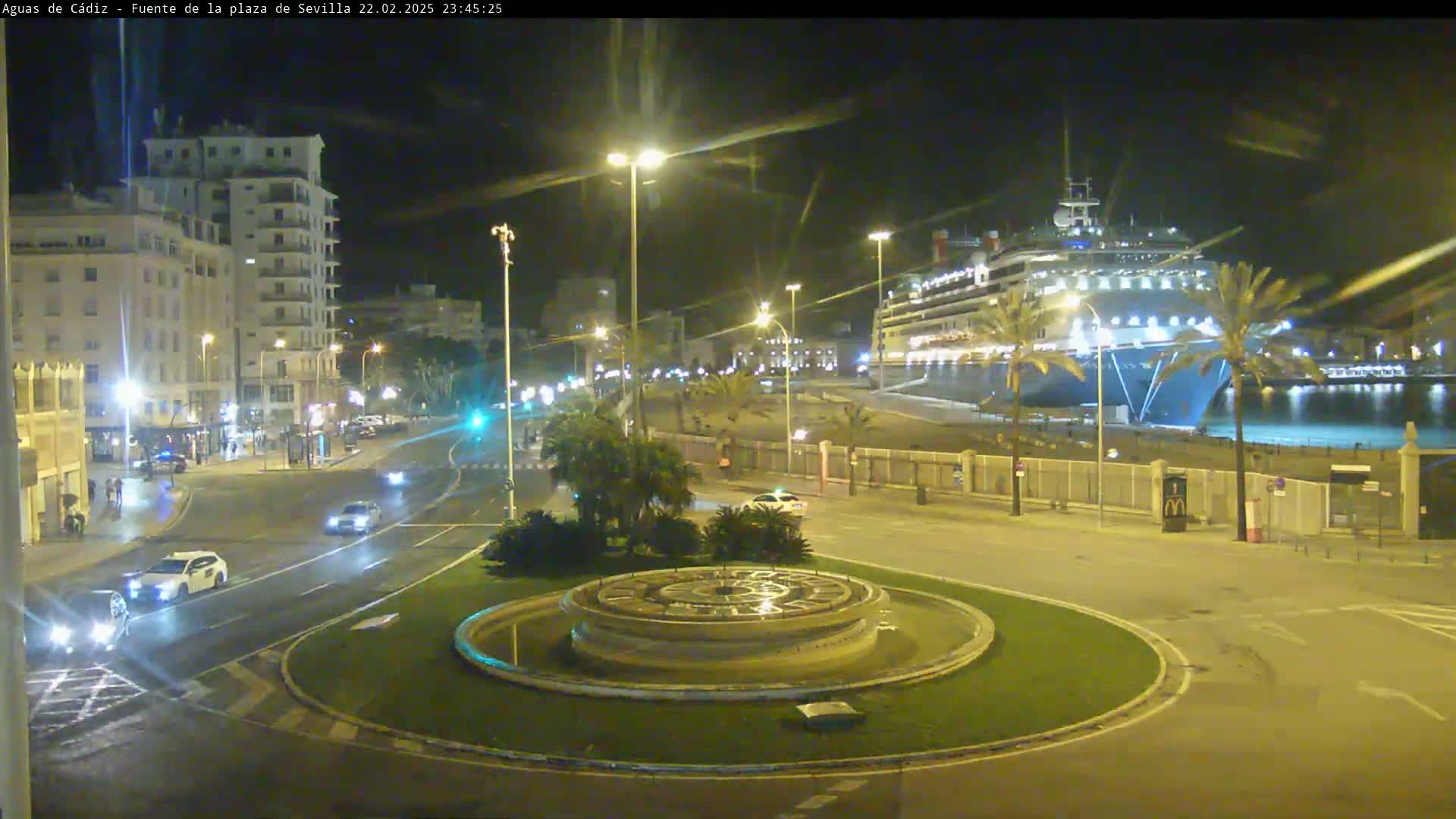A large cruise ship is docked at a port at night, with cars driving on a road that curves around a circular fountain in the foreground.