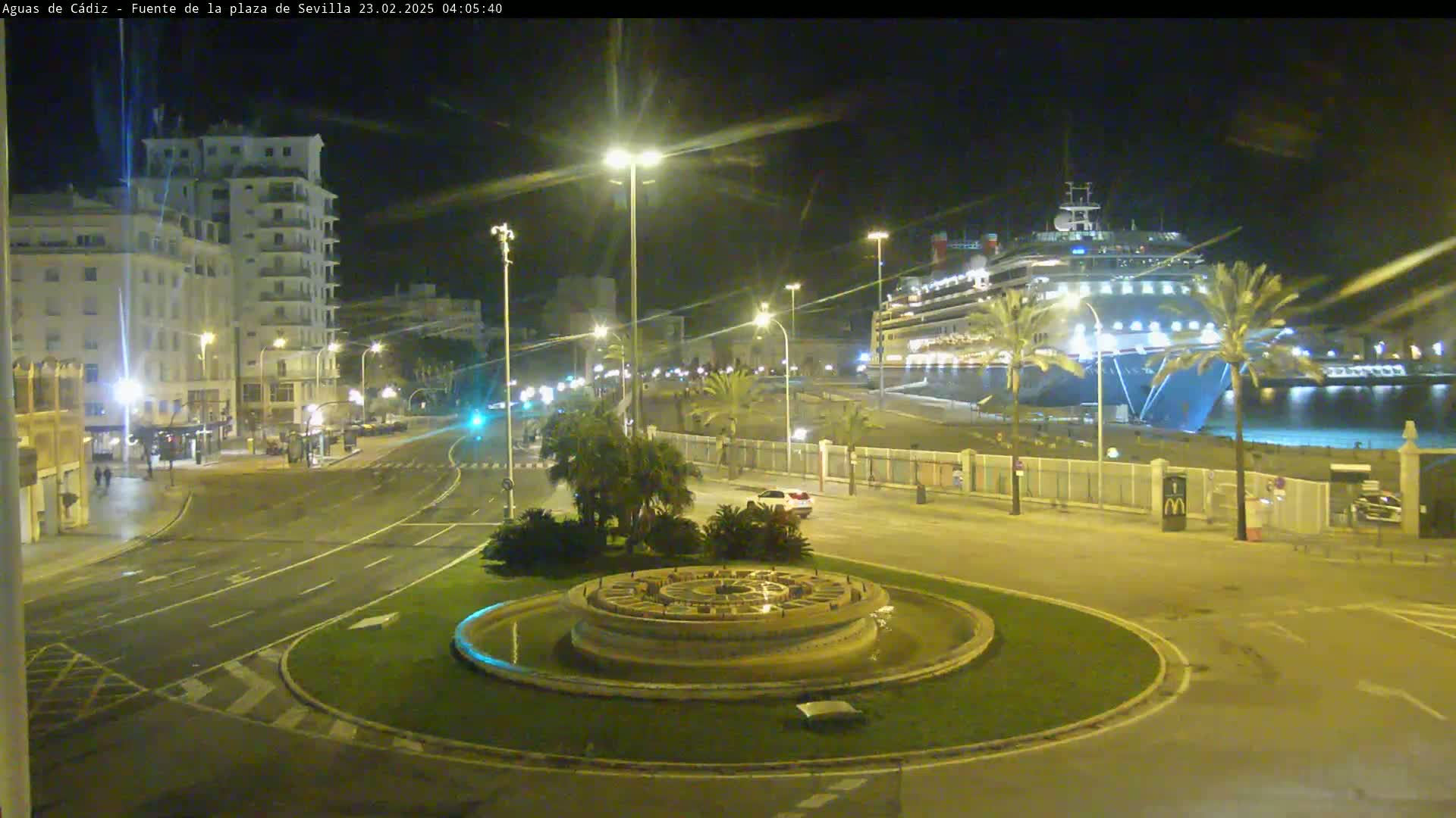 A large cruise ship is docked at a port at night, next to a circular fountain in a city square with buildings and streetlights illuminating the scene.