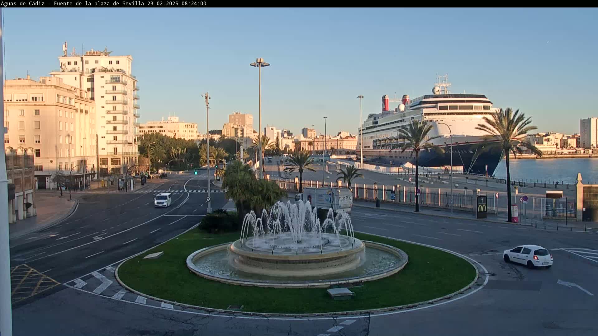 A large cruise ship is docked at a port city under a clear, bright sky, near a circular fountain and a roadway with sparse traffic.