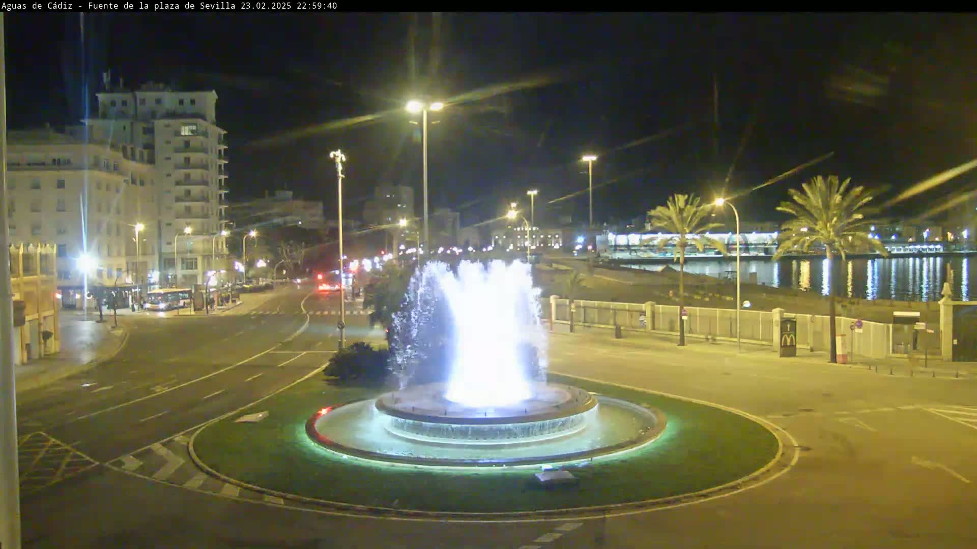 A brightly lit fountain in a roundabout is the center of a nighttime scene with city buildings and a calm body of water in the background.