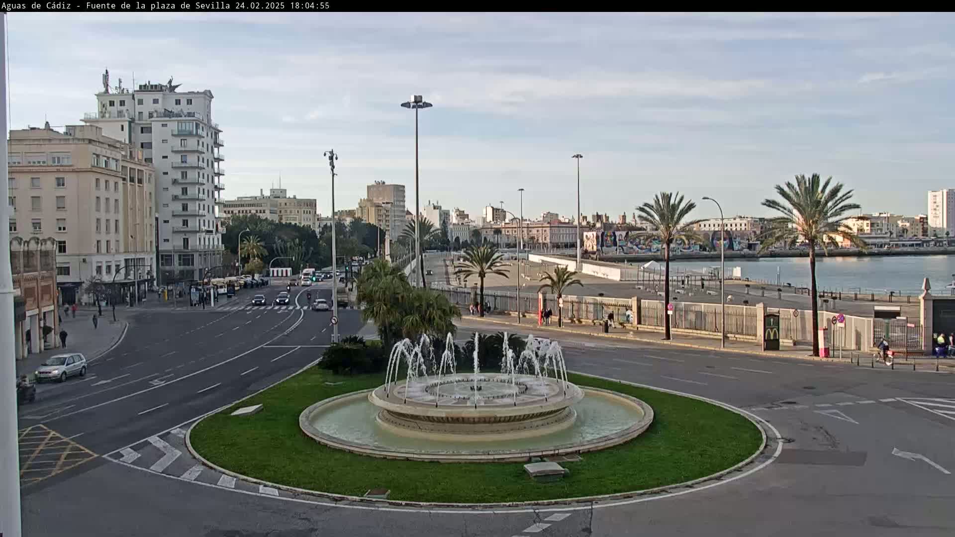 A city roundabout with a fountain in the center, surrounded by buildings, palm trees, and roads, overlooks a body of water under a partly cloudy sky.