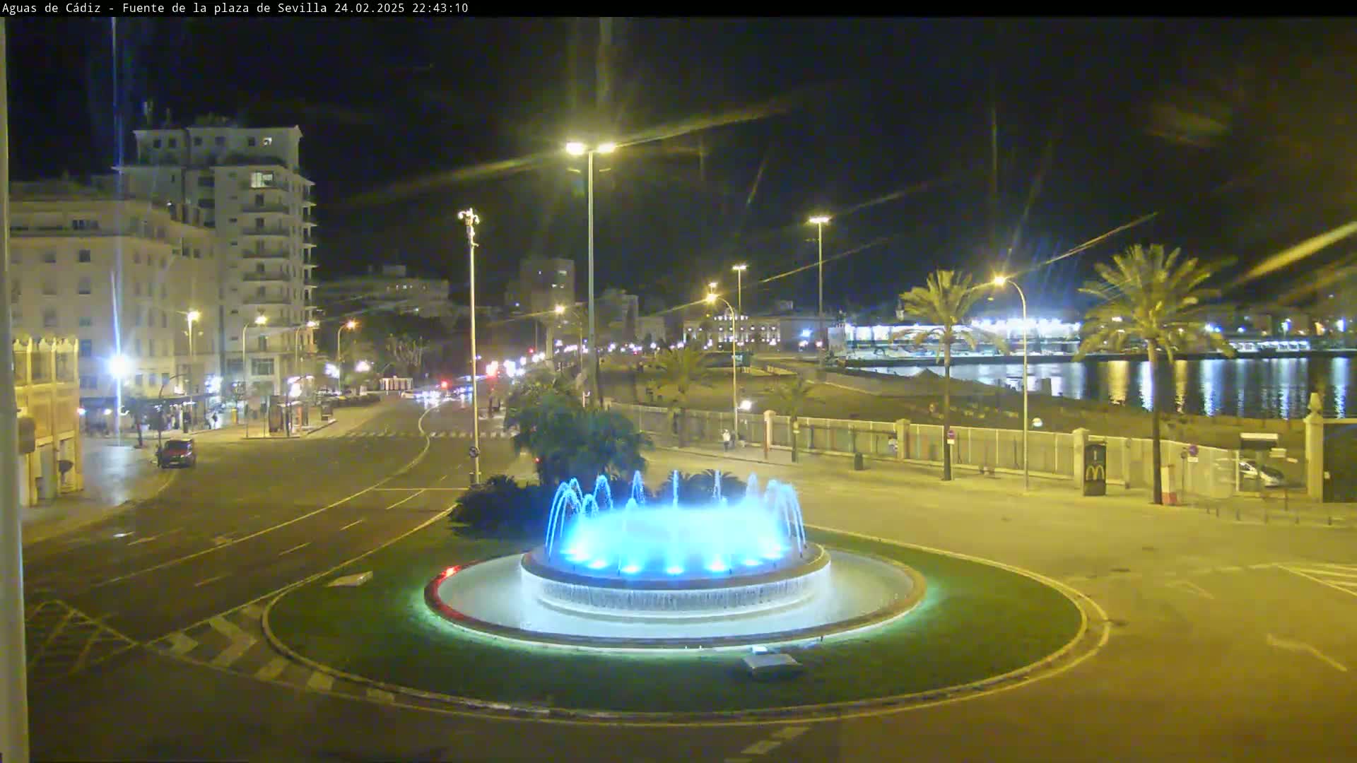 A nighttime view of a city square featuring a circular fountain with blue lights, illuminated streets, buildings, and palm trees, near a body of water under clear skies.