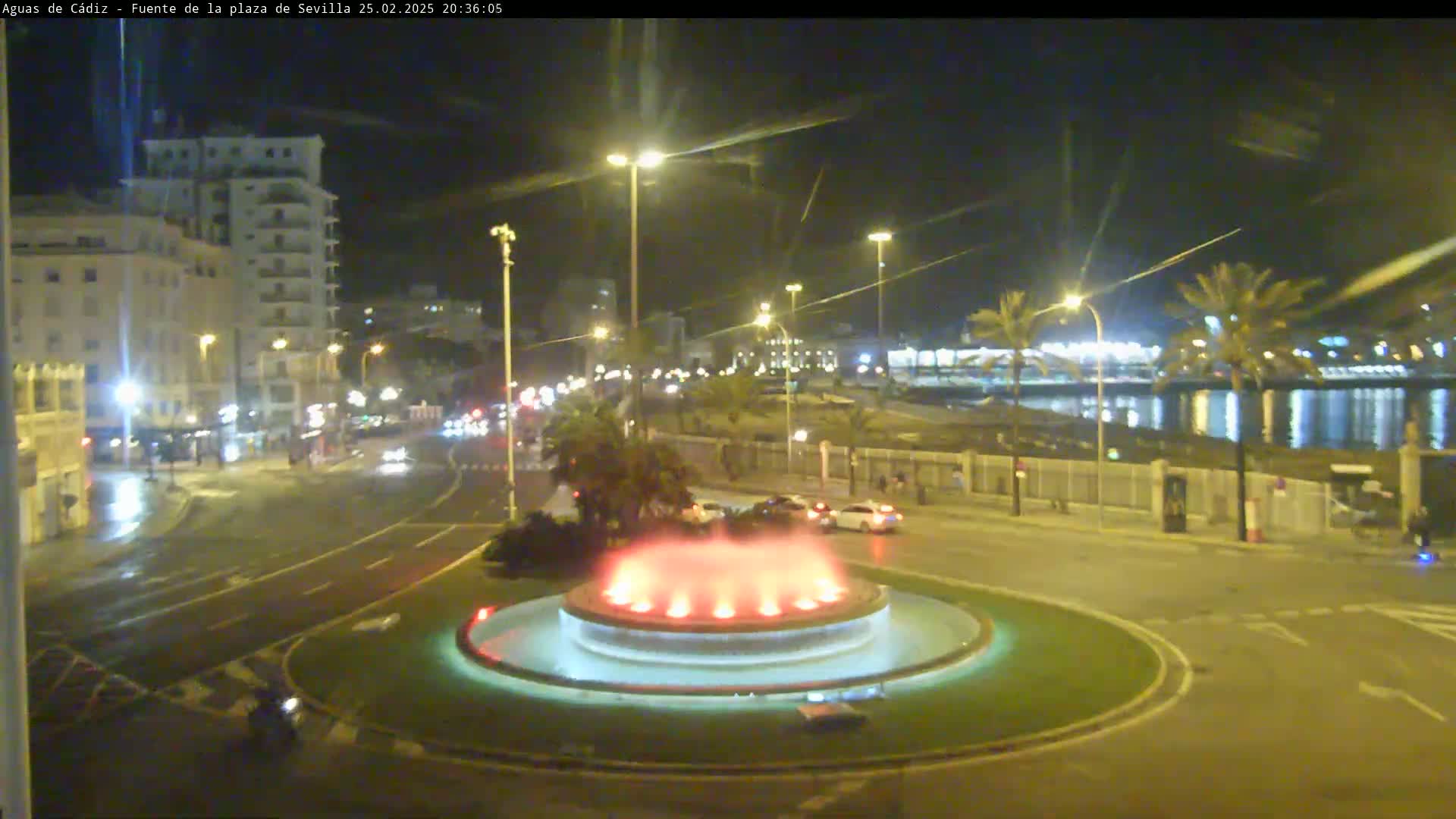 A nighttime view of a city roundabout with a lit fountain in the center, wet roads, and palm trees near a waterfront under overcast skies.