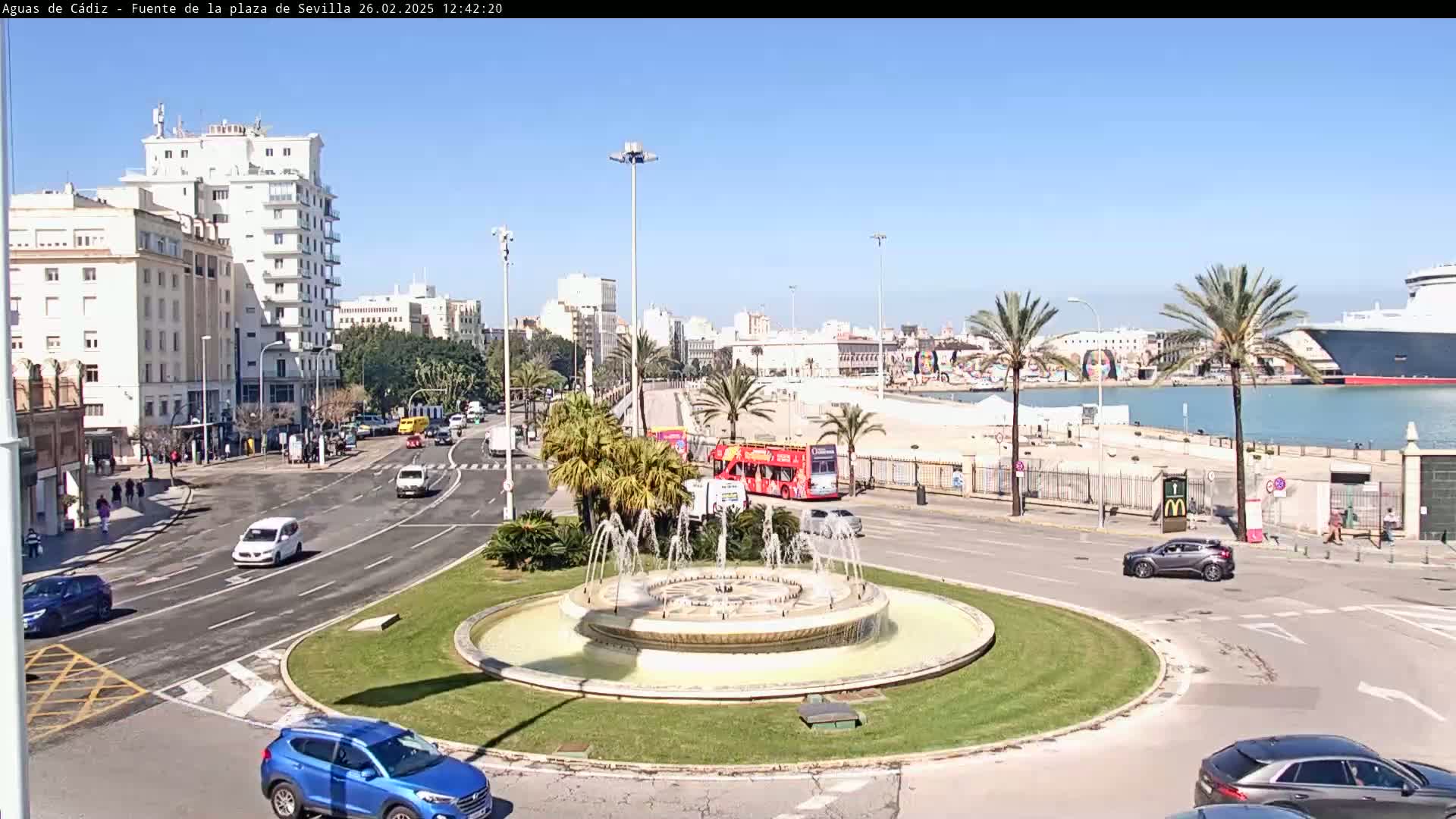 A traffic circle with a fountain in the center features several cars, a double-decker bus, and buildings, all under a sunny sky, with a harbor and cruise ship visible in the background.