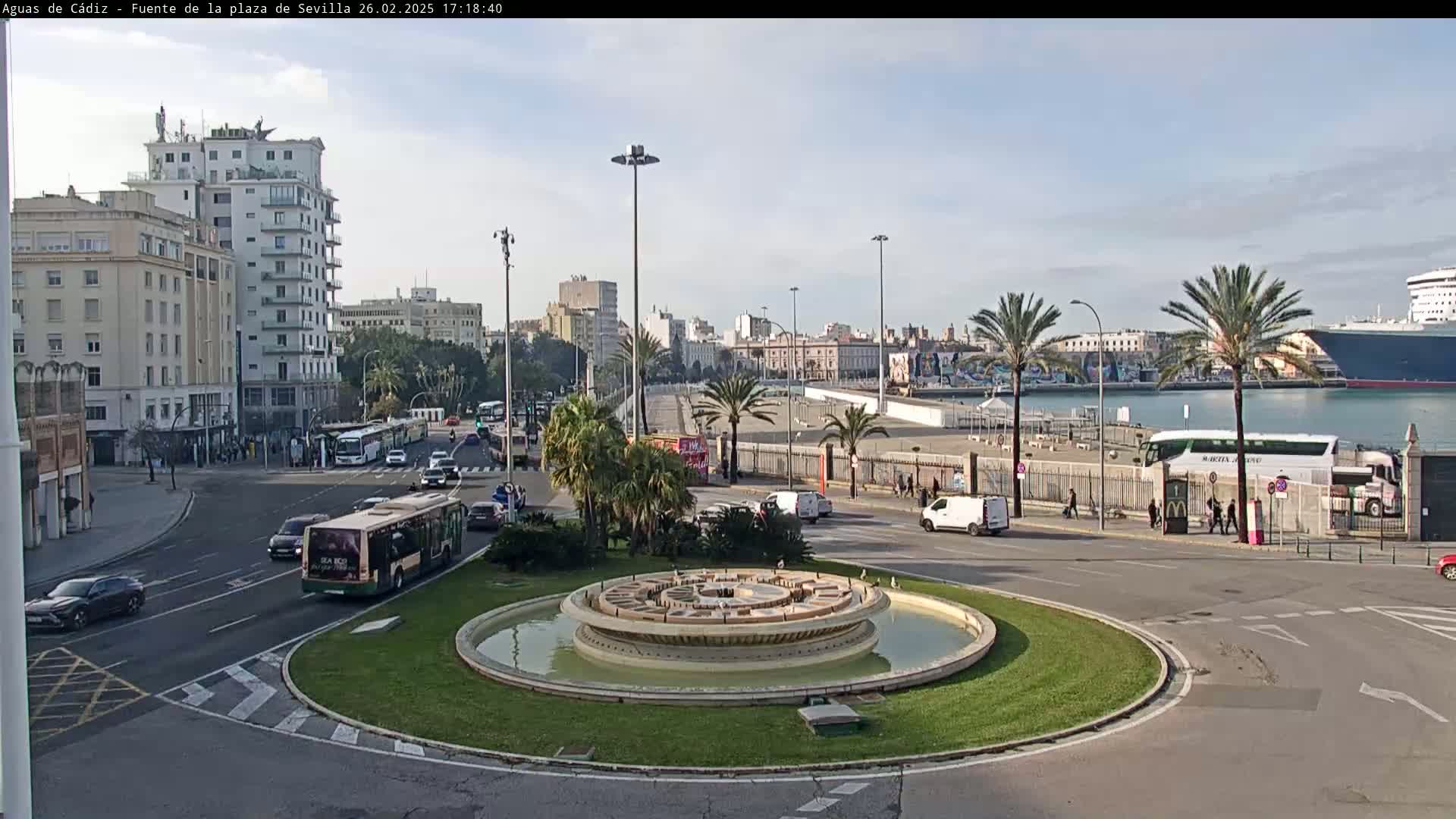 A circular fountain sits in a traffic circle in a city, with buildings, buses, and cars visible, and a large cruise ship in the harbor under a mostly clear sky.