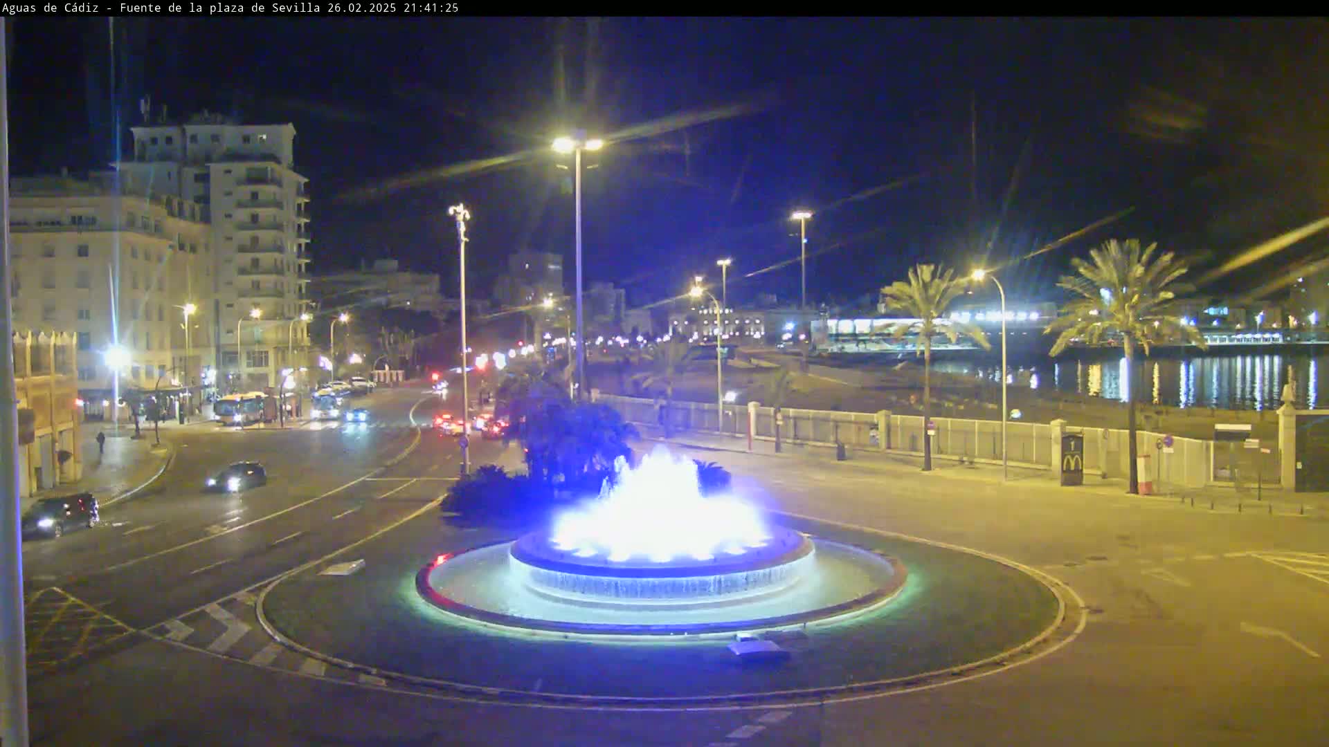A nighttime view of a city square with a circular fountain illuminated in blue, surrounded by roadways with cars and buildings, overlooking a calm body of water under a clear night sky.