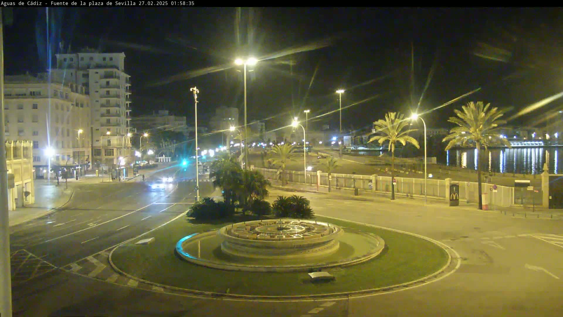 A nighttime view of a circular fountain in a city square, with a roadway and buildings visible under bright streetlights, and palm trees near a waterfront in the background.