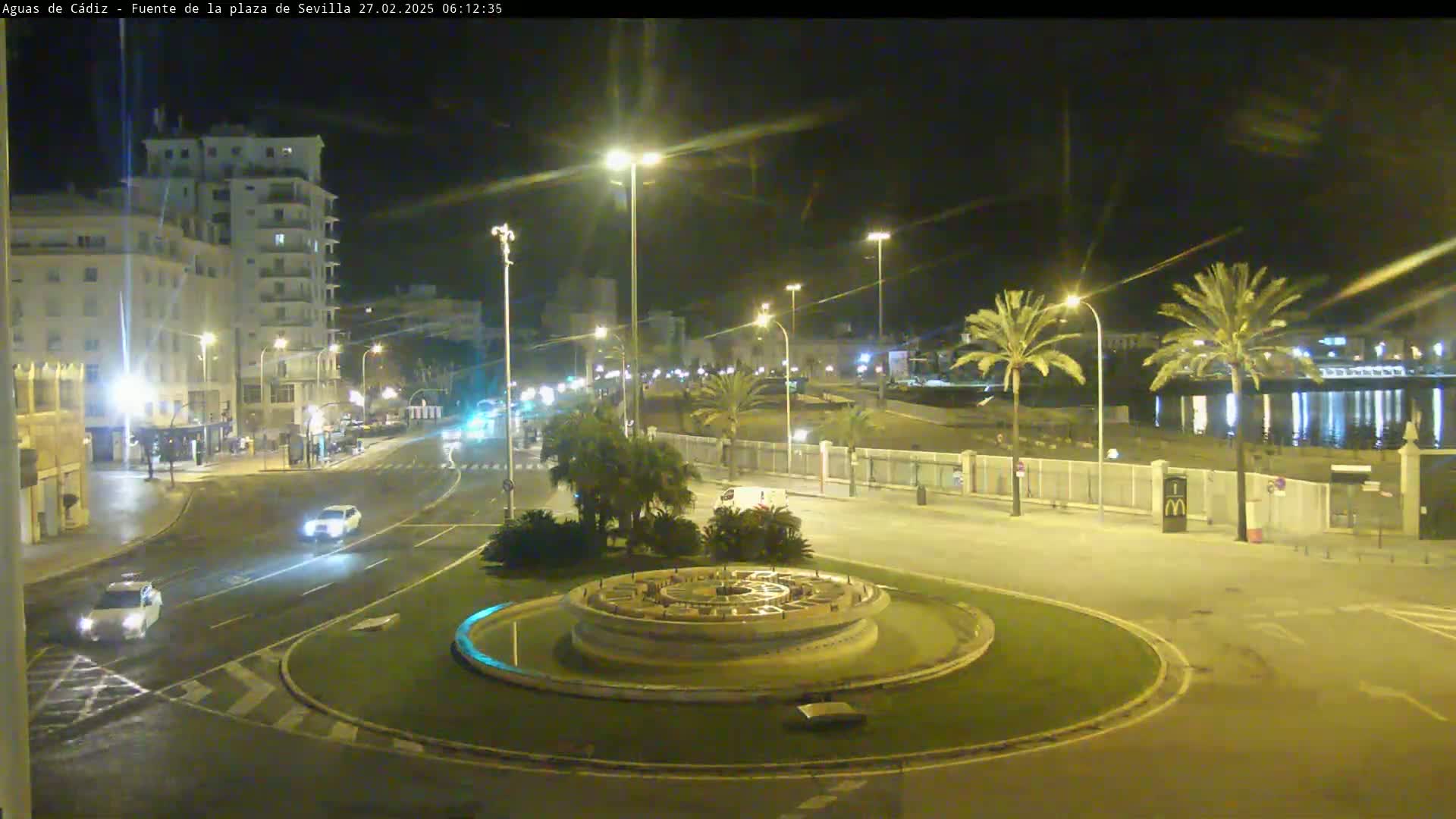 A nighttime view of a circular plaza with a fountain at its center, surrounded by streets with cars, palm trees, and buildings under well-lit conditions.